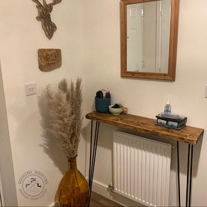 May include: A rustic wooden console table with black hairpin legs. The table is decorated with a vase of dried pampas grass, a small blue bowl, and a hand sanitiser bottle. The table is in a hallway with a white wall and a mirror on the wall.