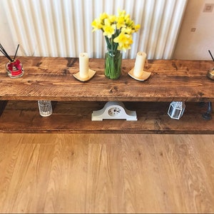 May include: A rustic wooden coffee table with a lower shelf. The table has a distressed brown finish and is decorated with candles, a clock, and a vase of yellow daffodils.