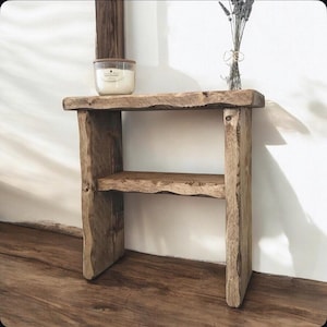 May include: A two-tiered wooden side table with a natural, unfinished look. A lit candle in a glass jar sits on the top shelf, and a bundle of dried lavender is placed on the right side. The table is made of rough-hewn wood, with a rustic aesthetic.
