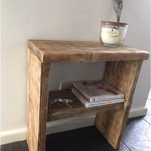 May include: A wooden side table with a shelf, made from light brown wood. A glass candle with a wooden lid and dried lavender sits on top. The shelf holds books and a metal figurine. The table has a rustic appearance.