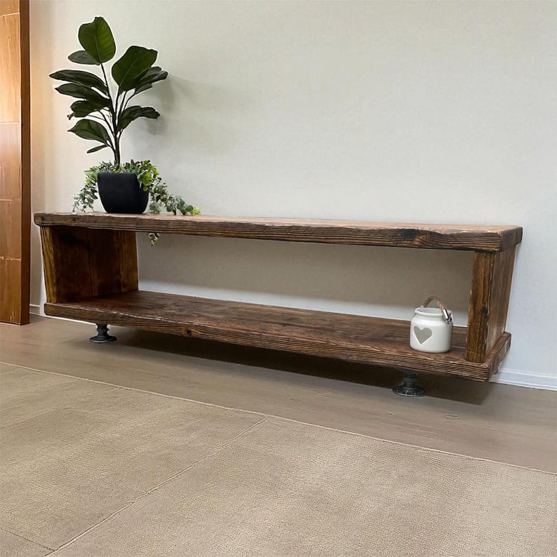 Puede incluir: Banco de madera r&uacute;stico, te&ntilde;ido oscuro, con un estante inferior y laterales angulados. El banco est&aacute; soportado por patas met&aacute;licas. Un peque&ntilde;o tarro blanco con un dise&ntilde;o de coraz&oacute;n se encuentra en el estante. Una planta en maceta con hojas verdes y una maceta negra est&aacute; encima.