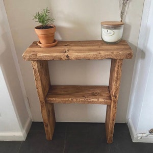May include: A rustic wooden shelf with two shelves. The shelf is stained a dark brown colour and has a distressed look. A small potted plant and a candle are on the top shelf.