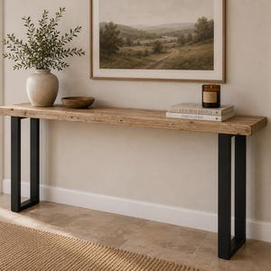 May include: A wooden console table with a light brown top and black metal legs. The table is decorated with a vase of greenery, a wooden bowl, books, and a candle. A framed landscape painting hangs above the table.