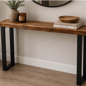 May include: A rustic wooden console table with a dark stained finish, supported by black metal legs. The table is decorated with a wooden bowl, books, a candle, and a vase with greenery. The table is approximately 122 cm long.