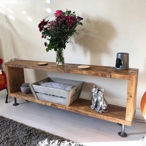 May include: A rustic wooden shelf with two shelves, featuring a gray wooden crate, two silver cat figurines, and a gray ceramic candle holder. The shelf is supported by industrial pipe legs.