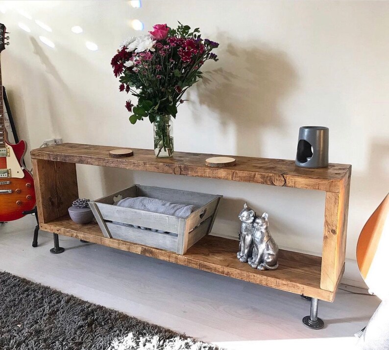 May include: A rustic wooden shelf with two shelves, featuring a gray wooden crate, two silver cat figurines, and a gray ceramic candle holder. The shelf is supported by industrial pipe legs.