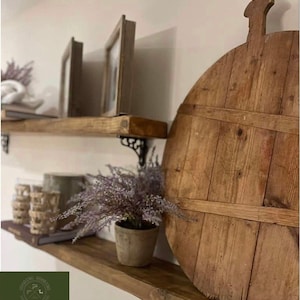 May include: Two wooden shelves with a rustic, farmhouse style. The top shelf has two framed pictures and a wooden round serving board with a handle on the bottom shelf. The shelves are supported by decorative metal brackets. The shelves are made of reclaimed wood and have a natural, unfinished look. The text "GOODDING WOODS" and "Cottage COLLECTION" is visible on the bottom shelf.