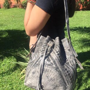 May include: A gray leather bucket bag with black embroidered details and a drawstring closure. The bag has a long strap and is worn over the shoulder.