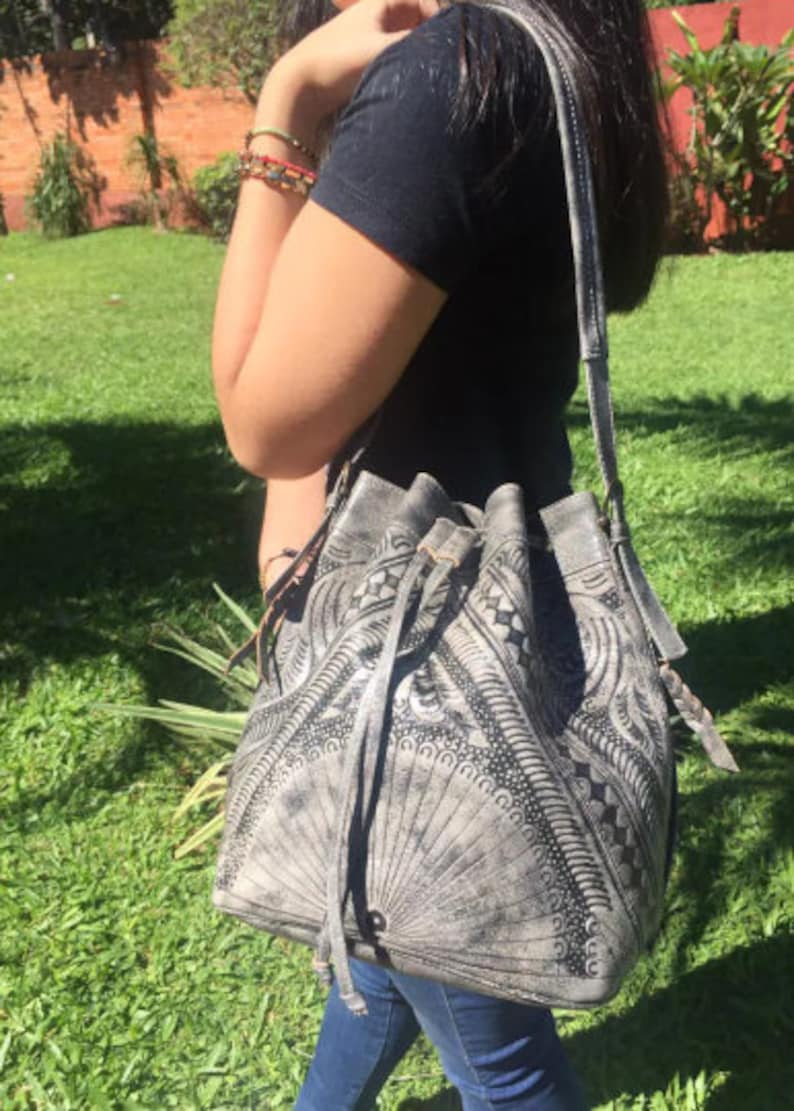 May include: A gray leather bucket bag with black embroidered details and a drawstring closure. The bag has a long strap and is worn over the shoulder.