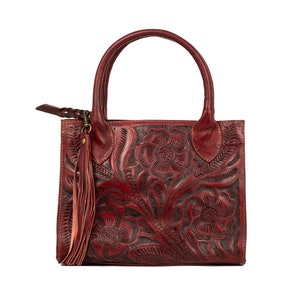 May include: A burgundy leather handbag with a floral embossed design. The bag features a top handle, a long tassel, and a zipper pull. The floral pattern is dark, creating a contrast with the burgundy leather. The bag is square-shaped.