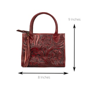 May include: A burgundy leather handbag with a floral embossed design. The bag has a top handle, a tassel, and measures 9 inches tall and 8 inches wide.