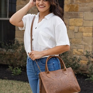 May include: A brown leather handbag with a floral embossed design. The bag has a shoulder strap and a tassel. The bag is carried by a person wearing a white shirt and blue jeans.
