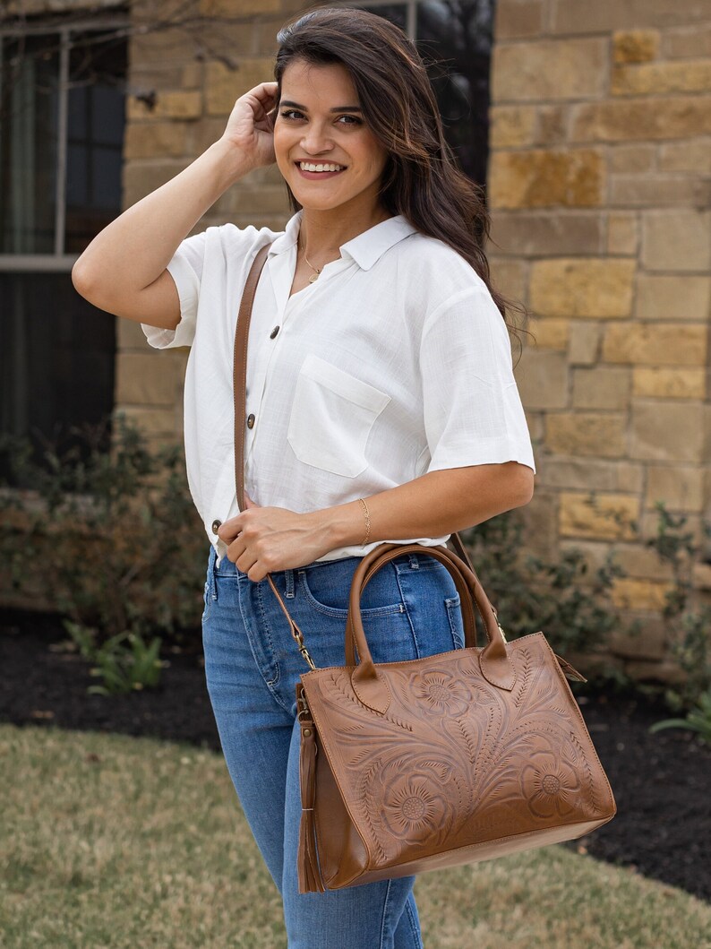 May include: A brown leather handbag with a floral embossed design. The bag has a shoulder strap and a tassel. The bag is carried by a person wearing a white shirt and blue jeans.