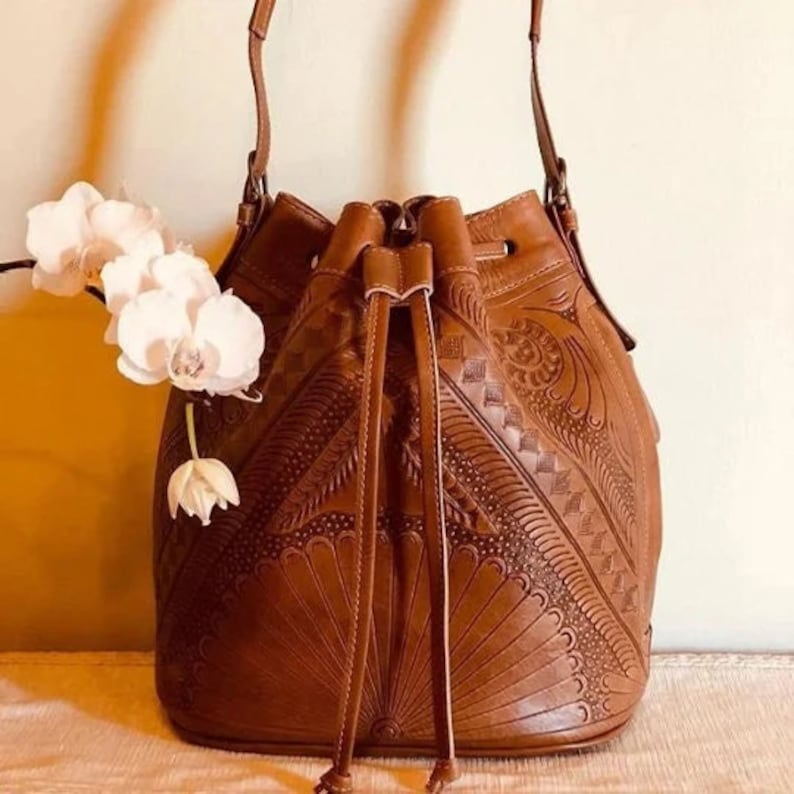 May include: A brown leather bucket bag with intricate embossed floral patterns. The bag has a drawstring closure and a long shoulder strap.