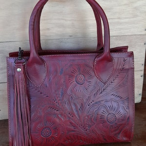 May include: Burgundy leather handbag with floral embossed design. Features dual handles, a detachable tassel, and a rectangular shape. The bag is made of a rich, deep red leather and has a classic, elegant style.