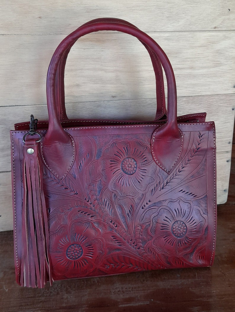 May include: Burgundy leather handbag with floral embossed design. Features dual handles, a detachable tassel, and a rectangular shape. The bag is made of a rich, deep red leather and has a classic, elegant style.