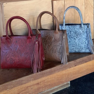 May include: Three leather tote bags in shades of red, brown, and gray. Each bag features a floral embossed design and a matching tassel. The bags have top handles and are displayed on a wooden shelf.