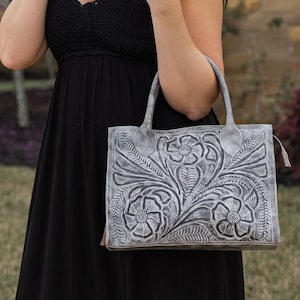 May include: A gray leather handbag with a floral embossed design. The bag features a rectangular shape with two handles and a tassel detail. The floral pattern includes detailed flowers and leaves.
