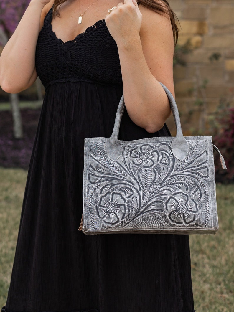 May include: A gray leather handbag with a floral embossed design. The bag features a rectangular shape with two handles and a tassel detail. The floral pattern includes detailed flowers and leaves.