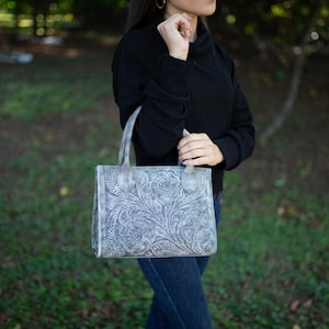 May include: A gray leather tote bag with a floral embossed design. The bag has two handles and a rectangular shape. The bag is held by a person wearing a black top and blue jeans.