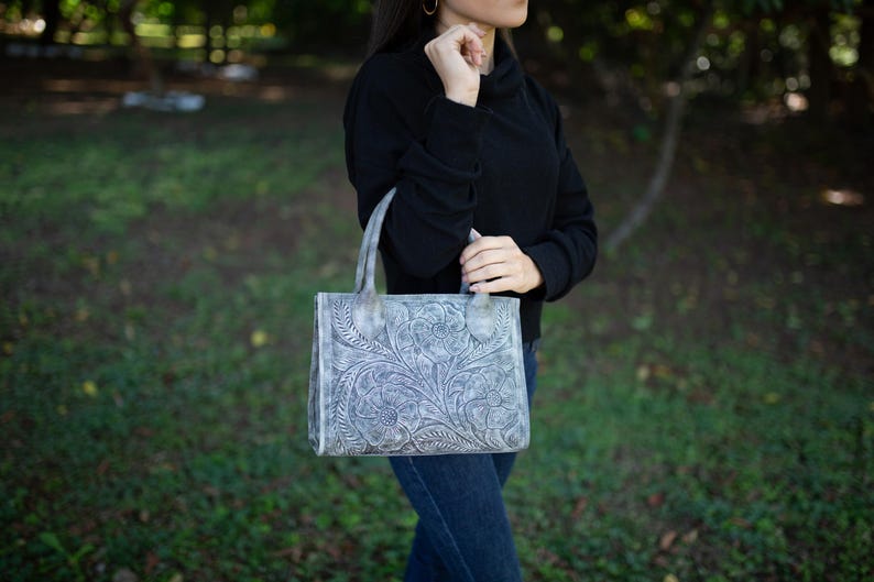 May include: A gray leather tote bag with a floral embossed design. The bag has two handles and a rectangular shape. The bag is held by a person wearing a black top and blue jeans.