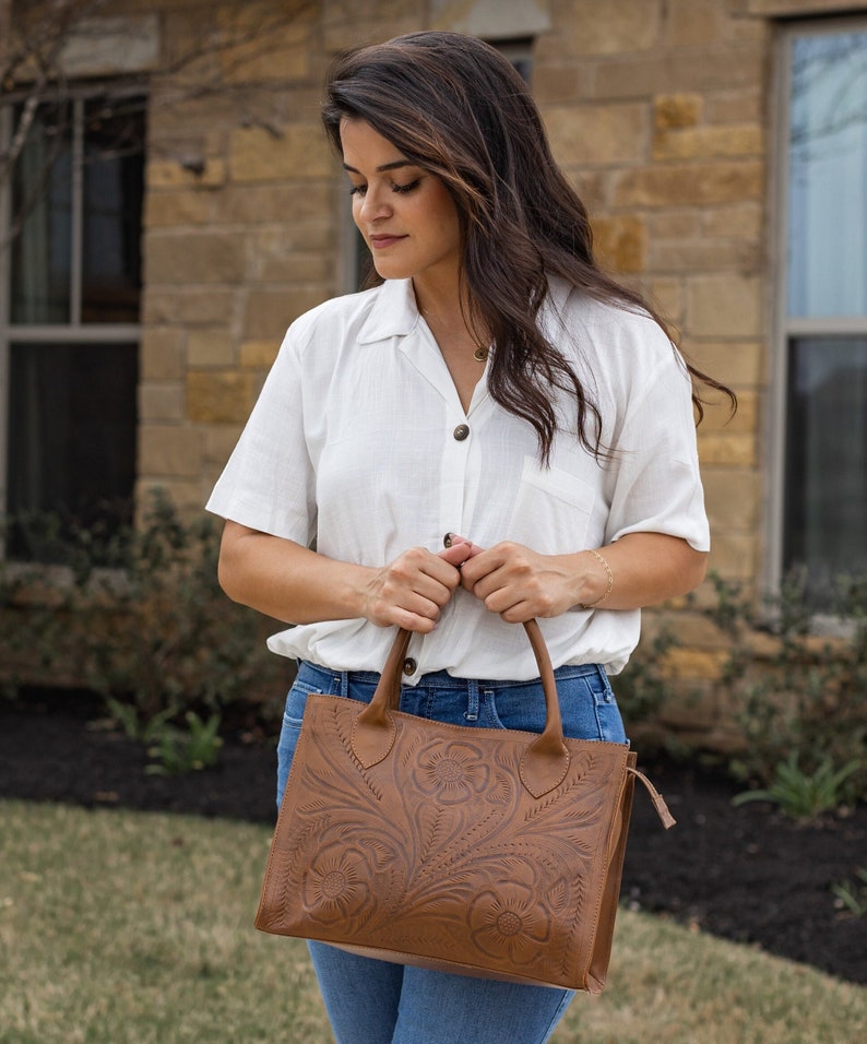 May include: A brown leather tote bag with a floral embossed design. The bag has two handles and a zipper closure. The woman is wearing a white button-down shirt and blue jeans.