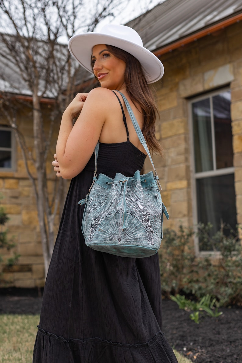 May include: A woman wearing a white hat and a black dress is carrying a teal blue leather bucket bag with a decorative pattern. The bag has a drawstring closure and a long strap.