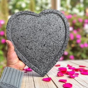 May include: A grey, heart-shaped decorative stone with a speckled texture and a raised border. The heart is held in a hand, with pink flower petals scattered on a wooden surface in the background. A unique home decor item.