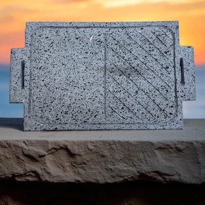 Puede incluir: Una tabla de servir rectangular gris moteado con una superficie texturizada y asas integradas. Una sección presenta ranuras diagonales. La tabla se muestra sobre una repisa de piedra contra un fondo de puesta de sol.