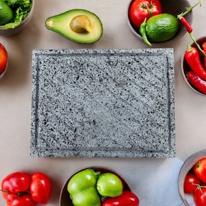 May include: A gray rectangular cutting board with a rough texture. The board is surrounded by bowls of fresh produce, including tomatoes, avocados, peppers, and limes.