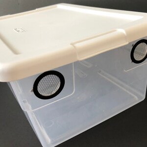 May include: A clear plastic storage container with a white lid. The container has two round holes with black mesh screens on the sides.