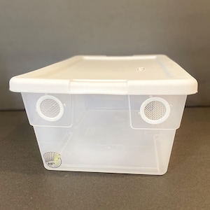 May include: A clear plastic storage container with a white lid. The container has two small, round, mesh vents on the sides. The container is empty.