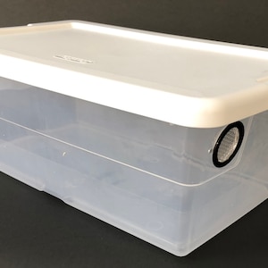 May include: A clear plastic storage container with a white lid. The lid has two round, black ventilation holes with mesh inserts.