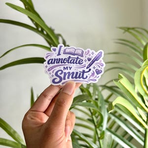 May include: A white sticker with the text "I annotate my smut" in purple script. The sticker features an open book, hearts, and a pen illustration. The sticker is held in front of a background of green leaves.
