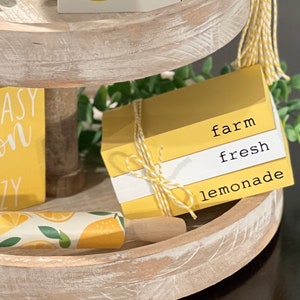 May include: A three-tiered wooden tray with a yellow and white lemon theme. The top tier has a sign that says "squeeze the day" with a lemon slice graphic. The middle tier has a sign that says "easy peasy lemon squeezy" and a box that says "farm fresh lemonade". The bottom tier has a rolling pin with a lemon print and a sprig of greenery.