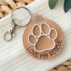 May include: A wooden pet tag with a white paw print design and the words "LOVE MY RESCUE" engraved around the edge. The tag is attached to a silver keyring with a clasp. The tag is round and made of light brown wood.