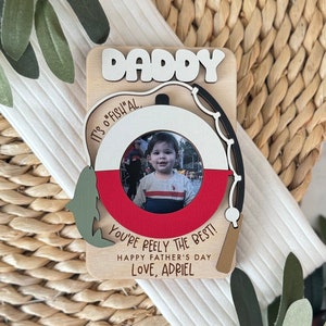 May include: Wooden Father's Day card with a photo frame shaped like a fishing bobber. The card says "Daddy, It's O "Fish" Al, You're Peely The Best! Happy Father's Day, Love, Adriel"