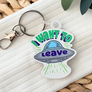 May include: A novelty keychain featuring a white, blue, and gray UFO design with the text "I WANT TO LEAVE". The keychain has a silver keyring and clasp.