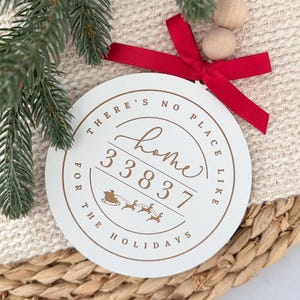 May include: A round, white Christmas ornament with the text "THERE'S NO PLACE LIKE HOME FOR THE HOLIDAYS" in brown. The ornament has a red ribbon and wooden beads for hanging. A sprig of evergreen and woven basket are in the background.