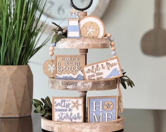 May include: A three-tiered wooden tray with a rustic whitewash finish. The top tier features a blue and white lighthouse, a wooden starfish, and a beaded garland. The middle tier has a sign that reads "meet me at the beach" and a sign that reads "wild & free just like the sea". The bottom tier has a sign that reads "salty kisses & starfish" and a sign that reads "home".
