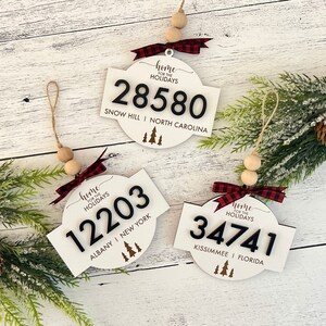 Puede incluir: Tres adornos navideños de madera blanca con números negros y el texto "Home for the Holidays". Cada uno tiene una cinta a cuadros roja y negra, cuentas de madera y una cuerda para colgar. Los adornos muestran diferentes códigos postales y nombres de ciudades.