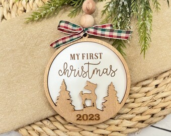 Op de afbeelding: Witte houten kerstbal met de tekst "Mijn eerste kerst" en een hert silhouet. Het jaar 2023 is ook op de kerstbal opgenomen.