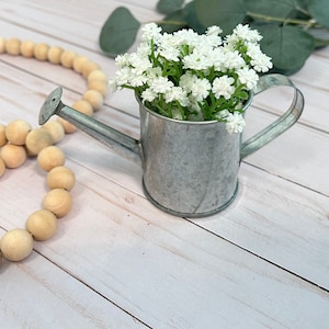 May include: A small, galvanized metal watering can with a white flower arrangement inside. The watering can is sitting on a white wooden surface.