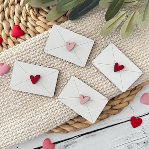 Pode incluir: Quatro decorações de envelopes de madeira branca com um pequeno detalhe de coração. Dois envelopes têm corações vermelhos e dois têm corações rosa. Pequenas decorações em forma de coração vermelhas e rosa estão espalhadas. Em uma superfície tecida.
