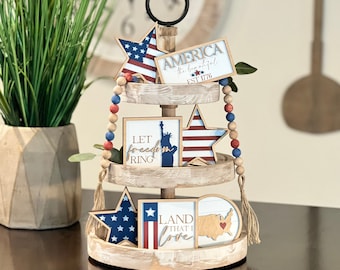 May include: A three-tiered wooden tray with a black handle, decorated with red, white, and blue stars, beads, and wooden signs. The signs feature patriotic phrases like "America the beautiful", "Let freedom ring", and "Land that I love".