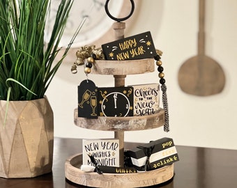 May include: A three-tiered wooden serving tray with a black metal ring handle. The tray is decorated with gold beads and features black and gold New Year's themed signs with phrases like "Happy New Year", "Cheers to the New Year", and "New Year Wishes Midnight".