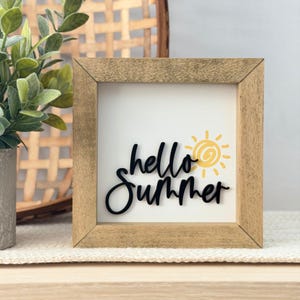 May include: A square wooden framed sign with the words "hello Summer" in black script. A yellow sun graphic is to the right of the word "Summer". The sign is on a white background.