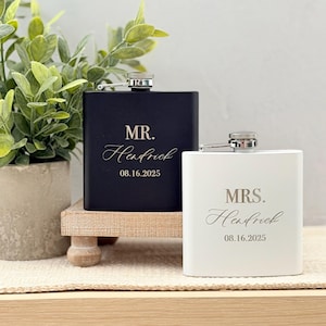 May include: Two personalised hip flasks, one black and one white, with silver detailing. The black flask reads "MR. Hendrick 08.16.2025" and the white flask reads "MRS. Hendrick 08.16.2025". They are displayed on a small wooden stand.