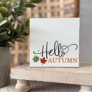 Puede incluir: Un letrero cuadrado de madera blanca con el texto Hello en cursiva negra y el texto AUTUMN en letras mayúsculas de color naranja. El diseño incluye una pequeña hoja de arce verde, una hoja de arce naranja y tres puntos dorados.