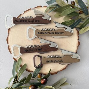 May include: Four wine bottle openers with brown leather handles and stainless steel components are displayed on a wooden surface. Each opener has a different engraved phrase: "Wire A LITTLE Laugh A LOT", "CAUTION: MAY CAUSE HONESTY", "THEY Whine 1 WINE", and "HAKUNA Moscato".